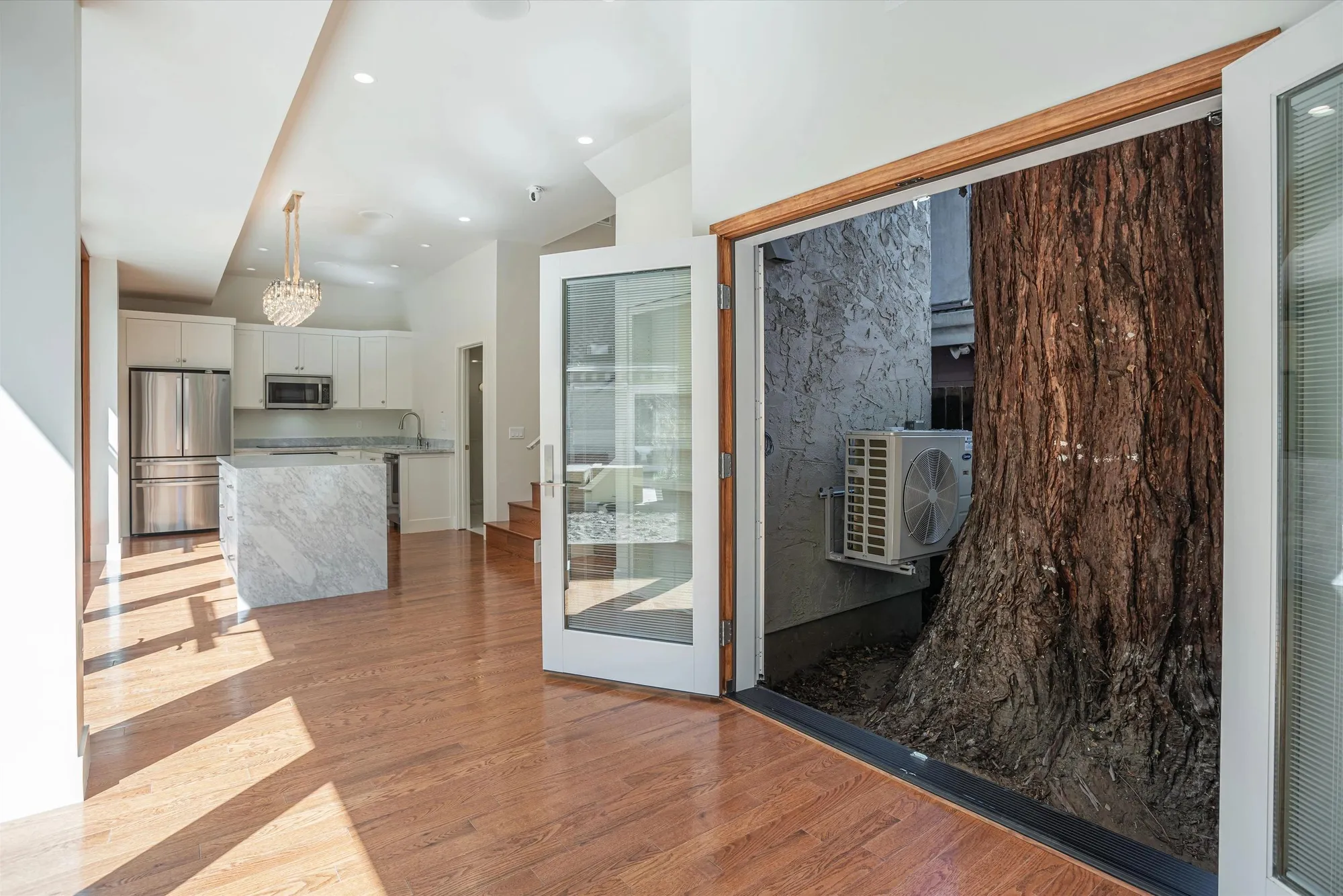 Open French doors beside a large tree trunk, with a kitchen visible beyond.