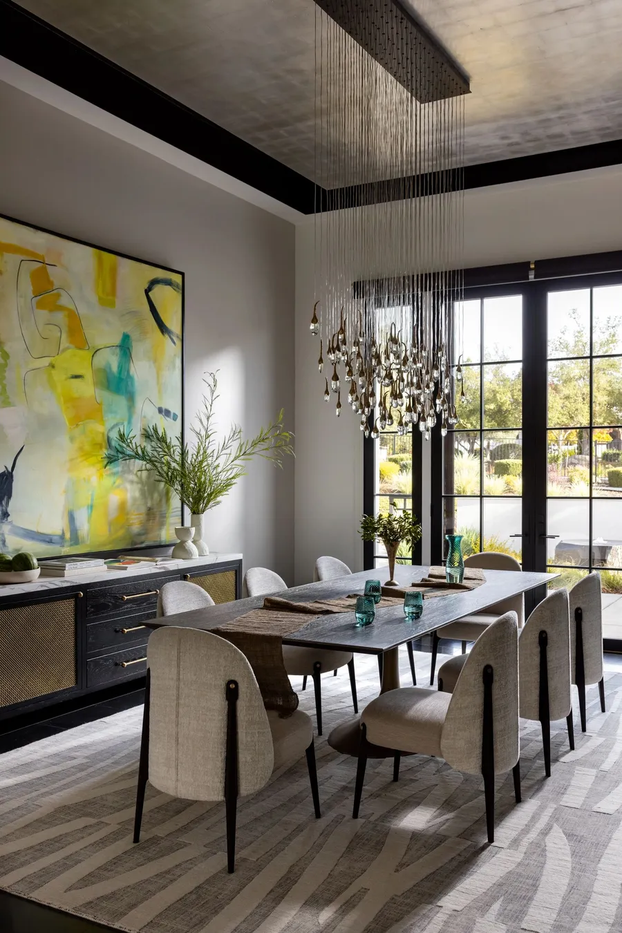 A dining room with a long table, teardrop chandelier, and glass doors to the garden.