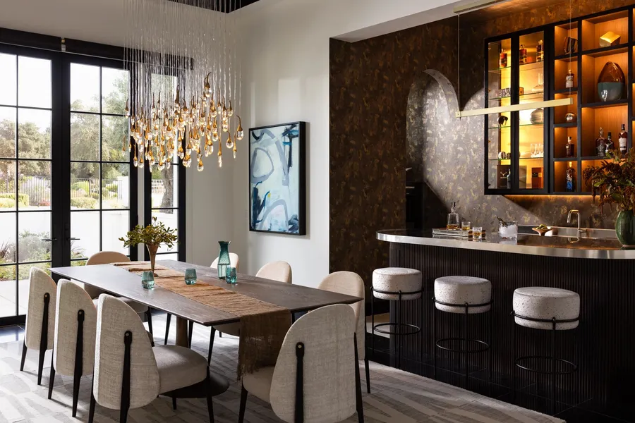 A dining table and custom bar with a glass chandelier and backlit cabinetry.