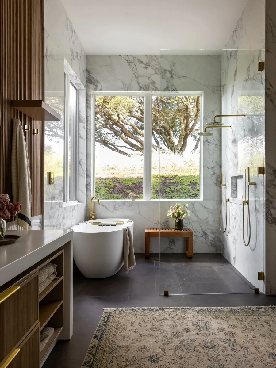 A bathroom with marble walls, a freestanding tub, and a window onto an oak tree.
